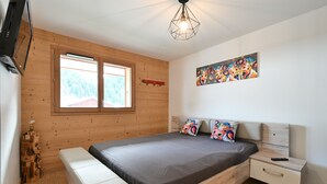A bedroom with a wooden ceiling, a bed with a grey mattress, a bedside table, a nightstand, a painting on the wall, and a window with a view of trees. - Apartment 'Le Barat'ski' with Mountain View, Private Terrace and Wi-Fi (Le Grand-Bornand)