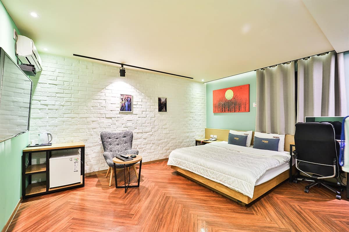 A hotel room with a brick wall, a bed, a desk with a computer, a chair, and a small kitchenette.