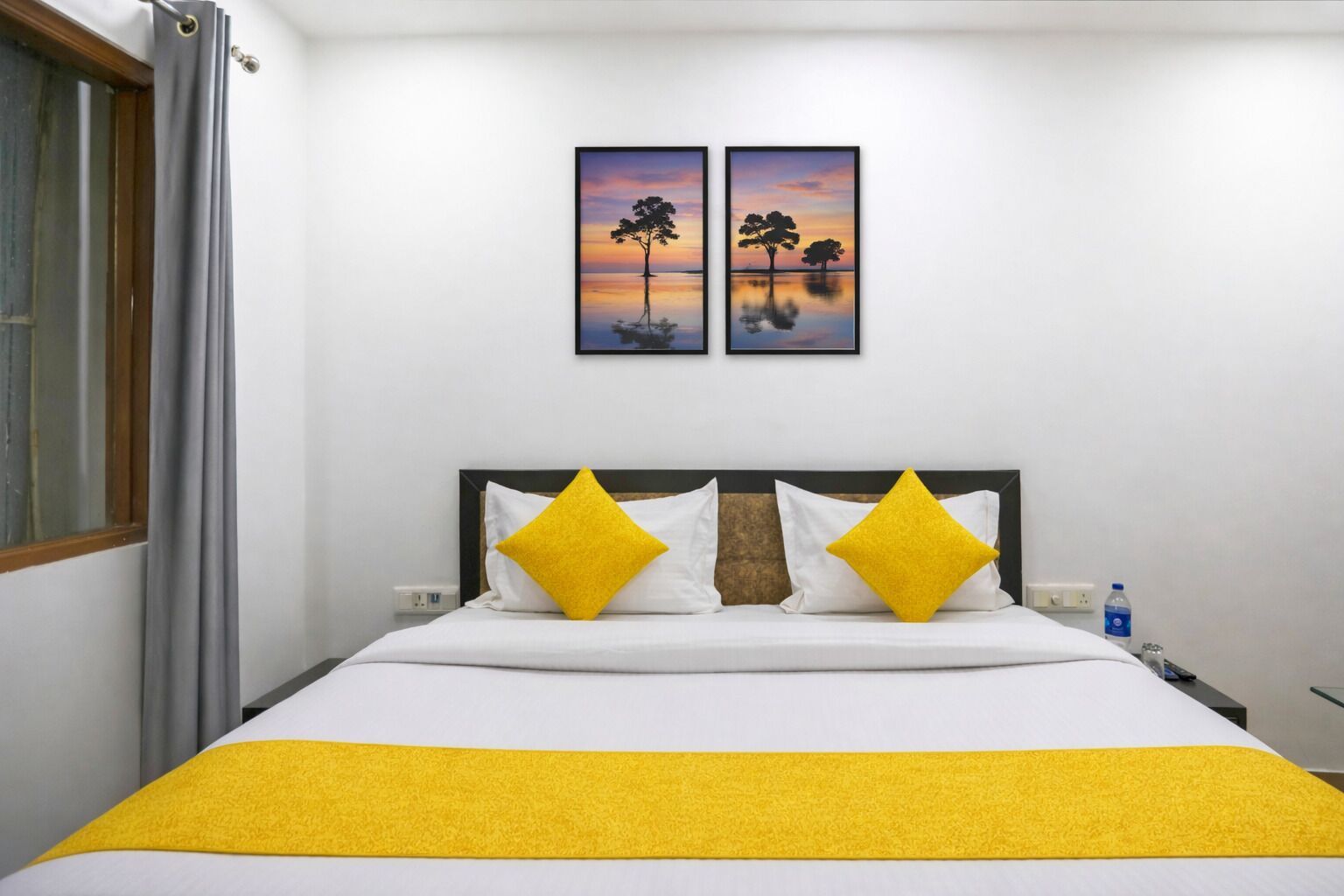 Een bed met wit beddengoed en twee gele kussens, een nachtkastje met een waterfles, en twee ingekaderde foto's aan de muur.