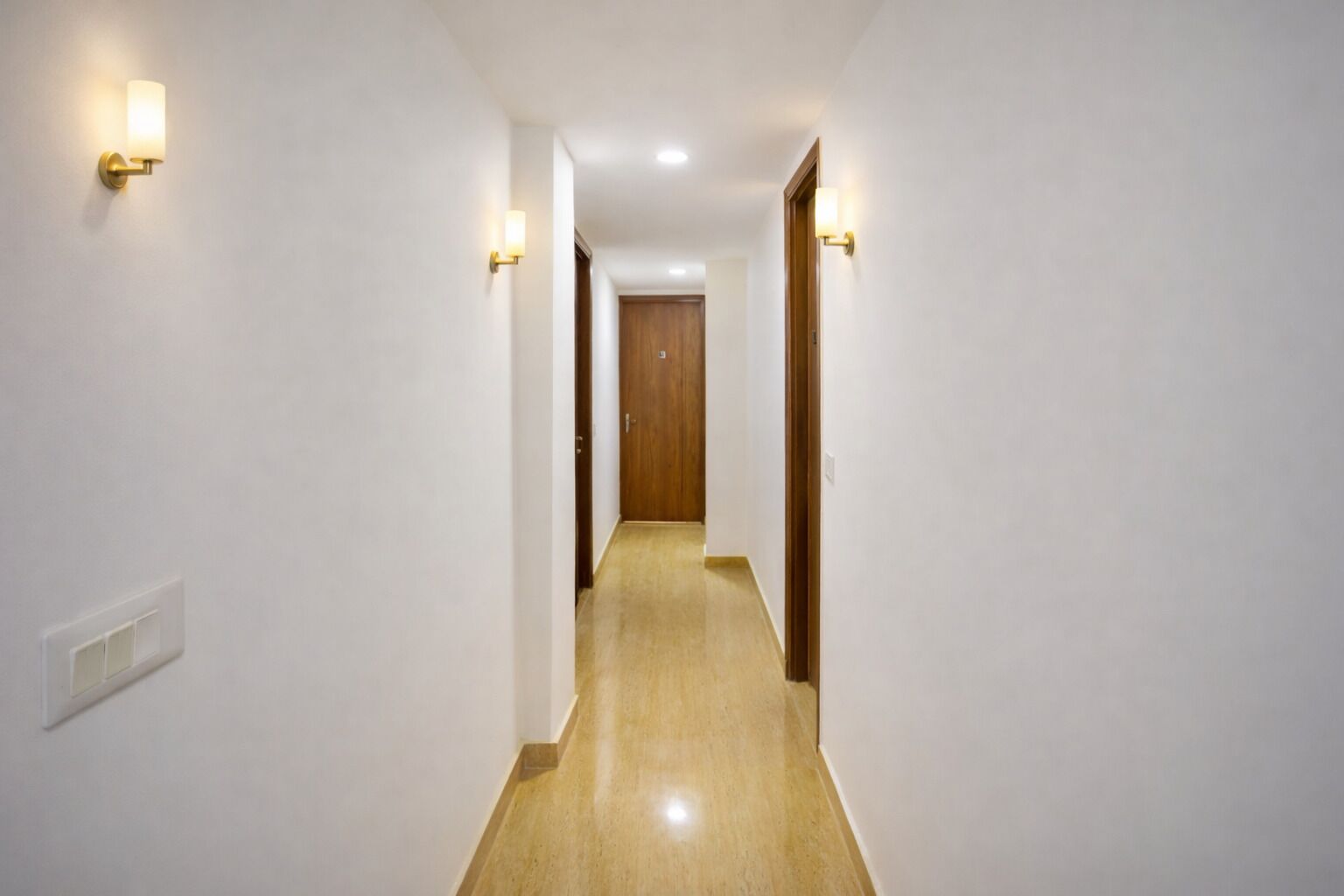 Un pasillo estrecho con suelo de madera, paredes blancas y dos lámparas fijadas a la pared.