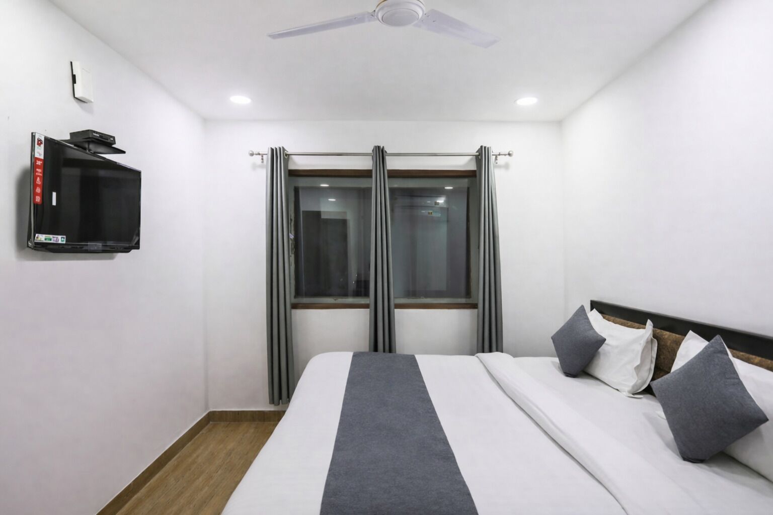 A hotel room with a bed, a TV mounted on the wall, a ceiling fan, and a window with curtains.