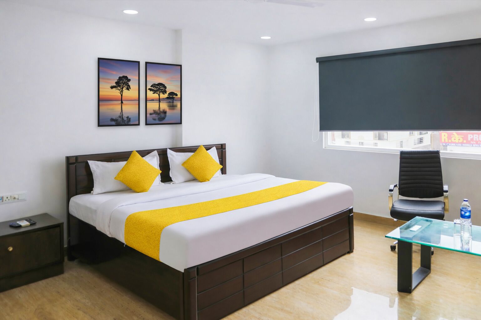 A hotel room with a bed, bedside table, chair, artwork, and a window with blinds.