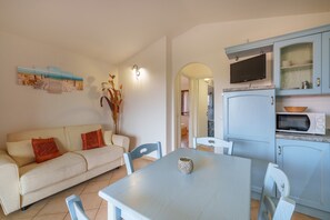 Uma sala de estar com sofá, cadeiras, mesa, televisão e uma área de cozinha com armários e eletrodomésticos.