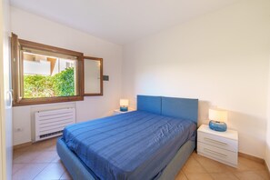Un dormitorio con cama, mesita de noche, lámpara, ventana y radiador.