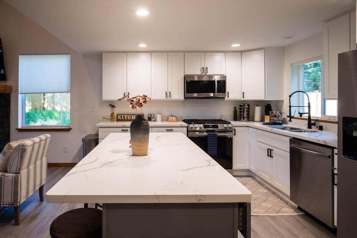 A modern kitchen with white cabinets, a central island, stainless steel appliances, and a view of greenery outside.