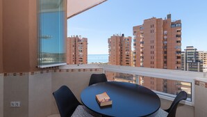 Een balkon met een tafel en stoelen, een boek op de tafel, en uitzicht op hoge gebouwen en de zee.