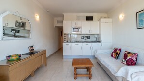 Een compacte leefruimte met een kitchenette, een bank, een houten tafel en een kastje met een decoratieve schaal.