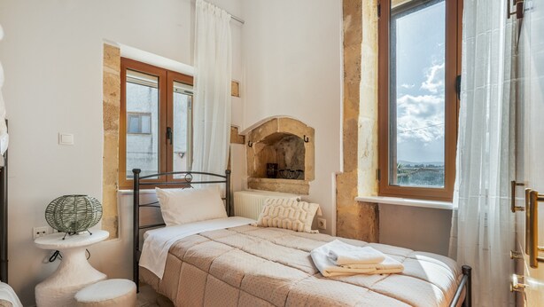 A bedroom with a bed, a bedside table, a small stool, a window with curtains, and a stone archway. - Palailimnos Apartment with Sea View, Wi-Fi, and Air Conditioning (Palelimnos)