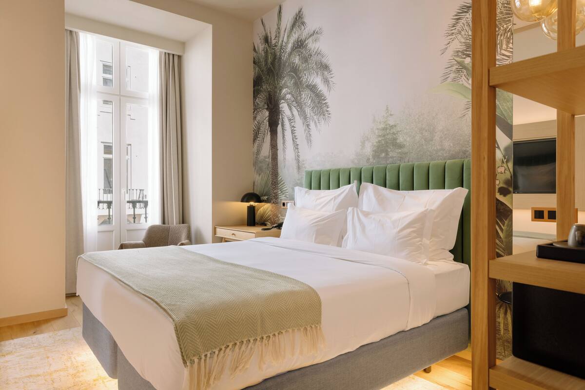 A neatly made bed with a green headboard, a large window with curtains, and a wall mural of a palm tree.