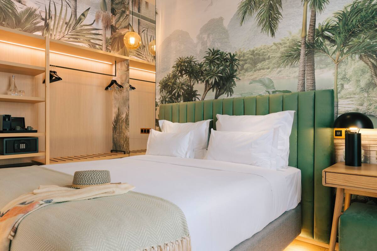A neatly made bed with white linens, a green upholstered headboard, and a wall mural depicting a tropical landscape.