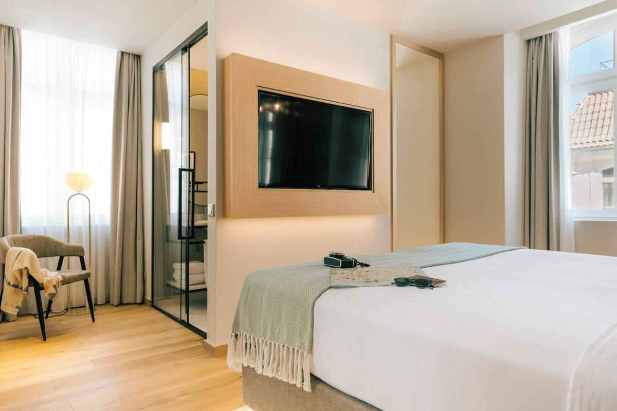 A modern hotel room with a large bed, a flat-screen TV mounted on the wall, a chair, and a floor lamp.