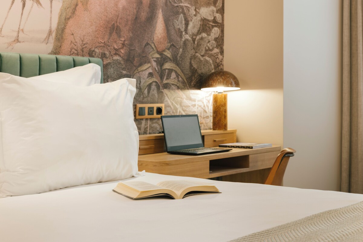 A hotel room with a bed, a desk with a laptop, a lamp, and a large wall painting.