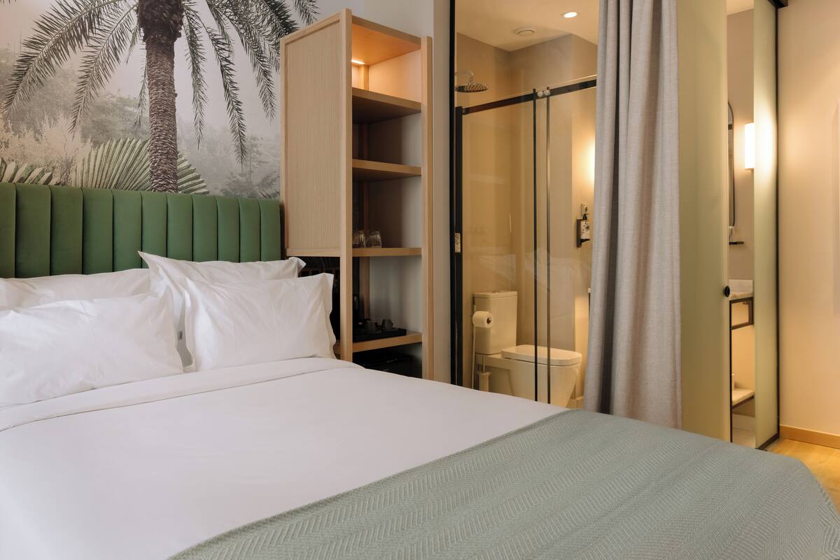 A hotel room with a bed, a green headboard, a wardrobe, a shower area, and a wall with a palm tree mural.