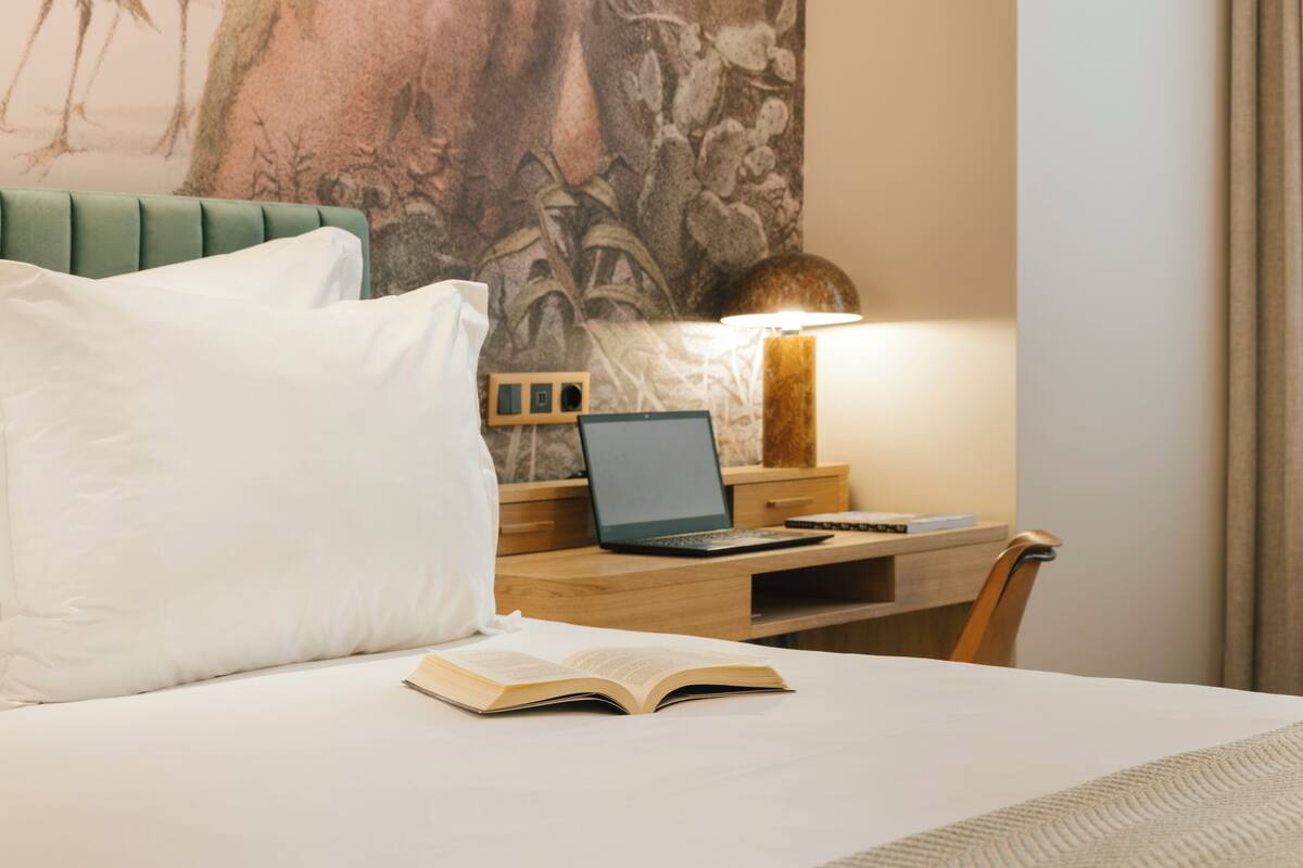 A hotel room with a bed, a desk with a laptop, a lamp, and a large wall painting.
