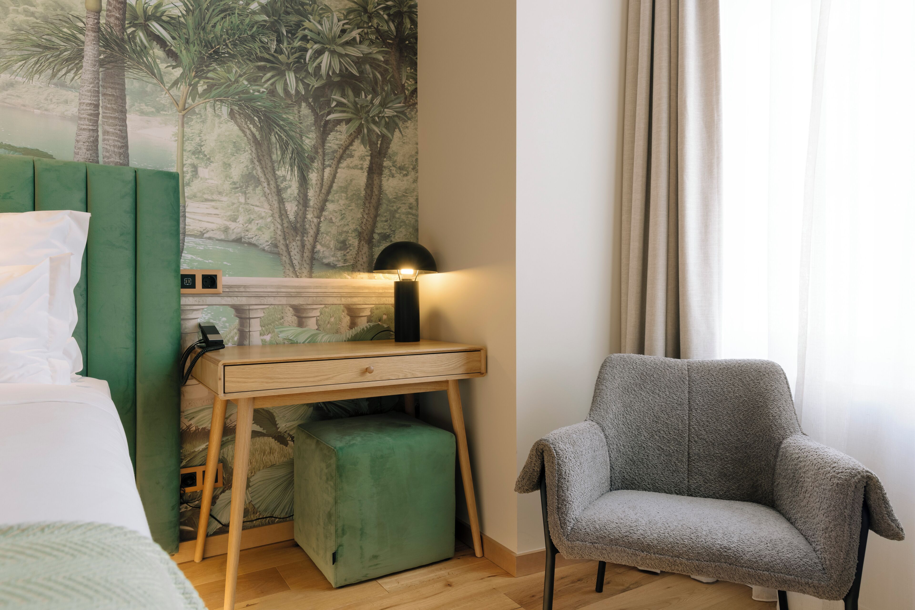 Une chambre à coucher avec un lit, un bureau avec une lampe, un pouf vert et un fauteuil gris.
