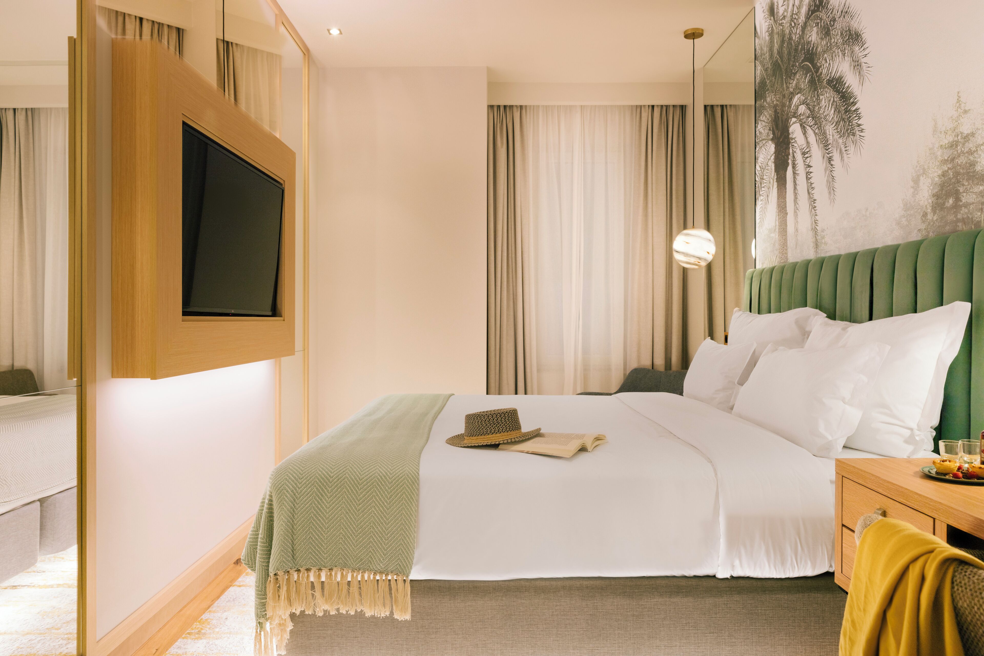 Quarto de hotel com cama, televisão na parede, mesa de cabeceira e uma janela grande com cortinas.