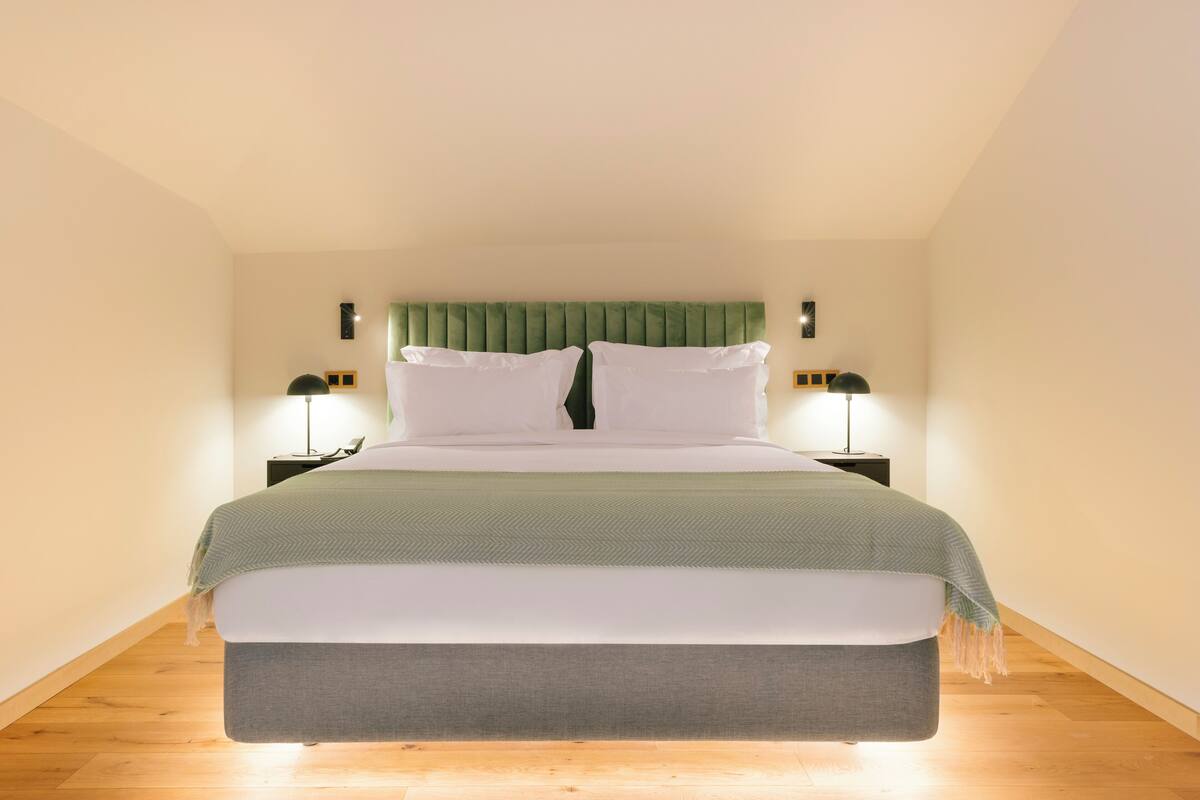 A modern bedroom with a large bed, green headboard, two bedside lamps, and a wooden floor.
