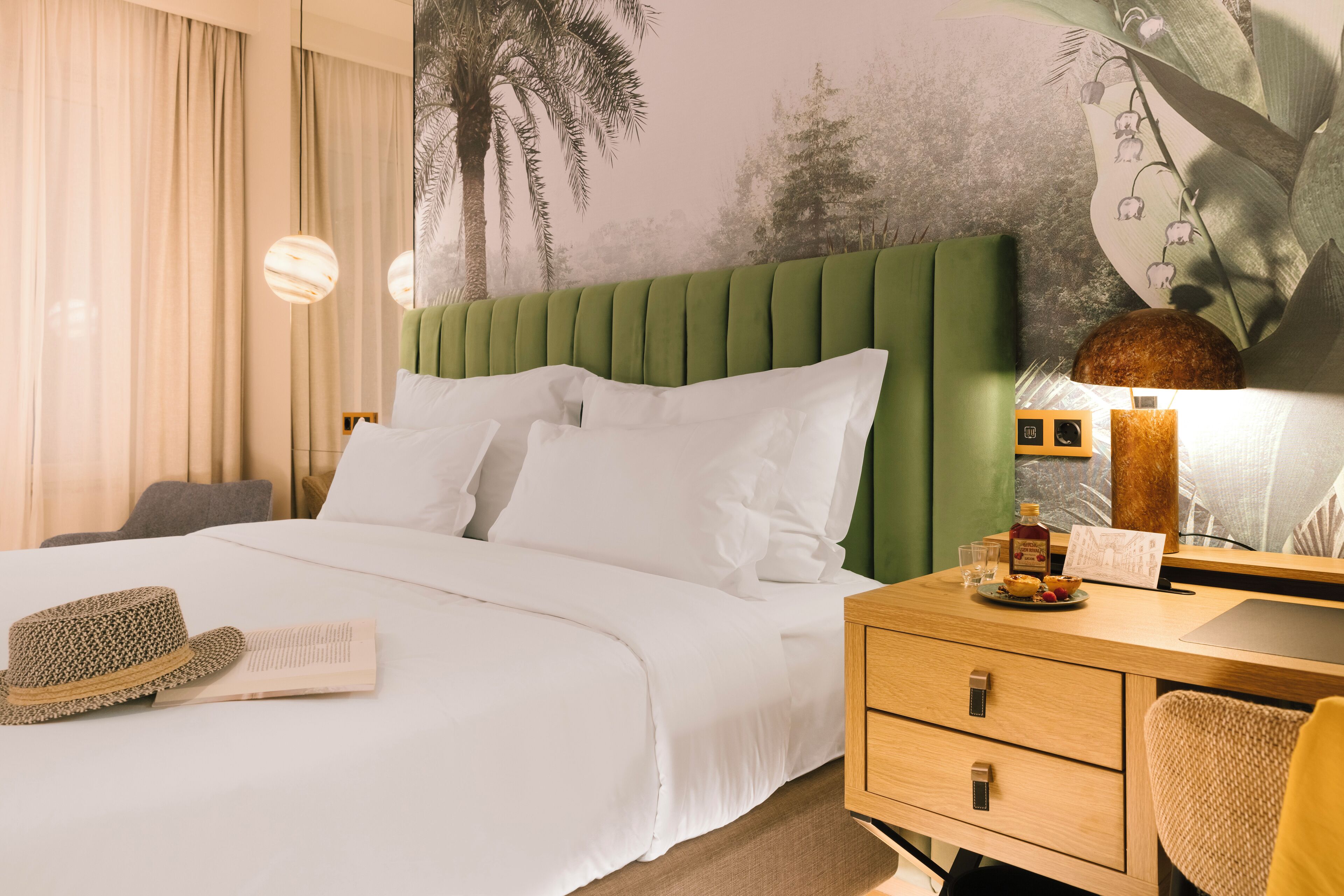 A hotel room with a green upholstered headboard, a white bedspread, a wooden desk with drawers, a lamp, and a wall mural.