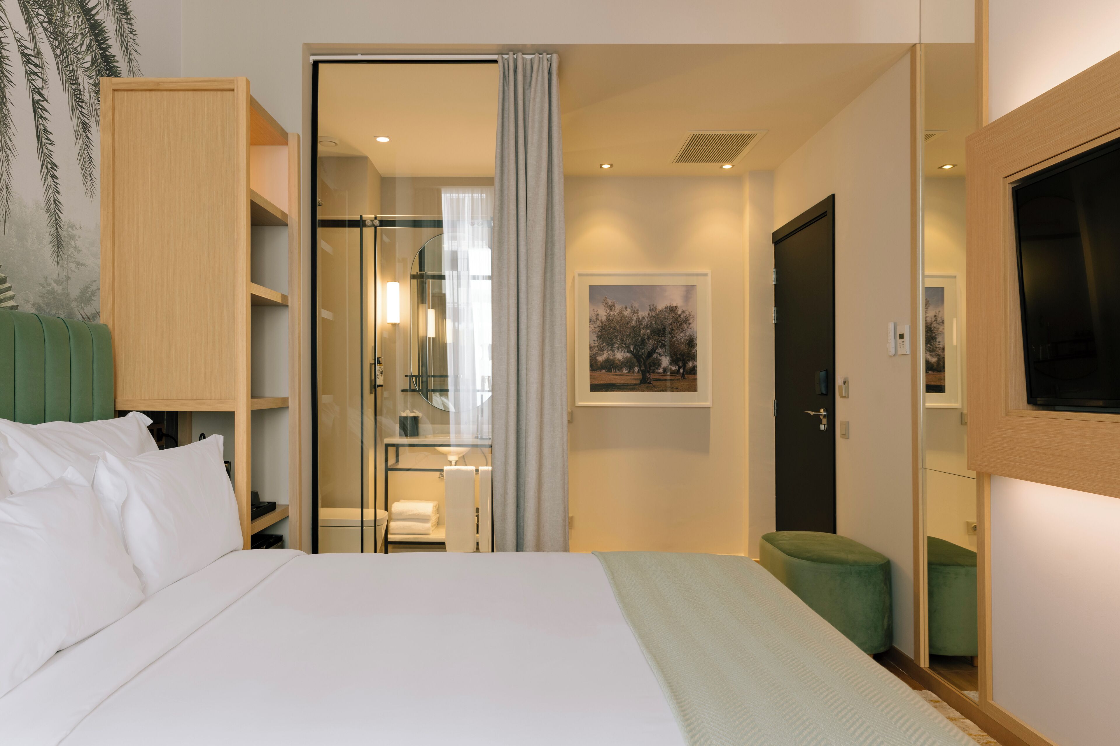 A hotel room with a bed, a green ottoman, a wardrobe, a bathroom with a glass shower, and a framed picture on the wall.