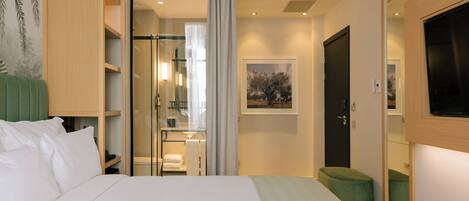 A hotel room with a bed, a green ottoman, a wardrobe, a bathroom with a glass shower, and a framed picture on the wall.
