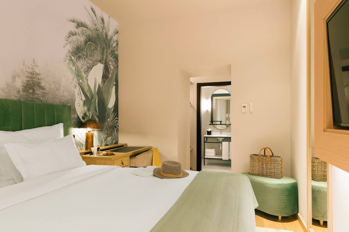 A hotel room with a bed, a nightstand, a lamp, a green ottoman, and a bathroom visible through an open door.