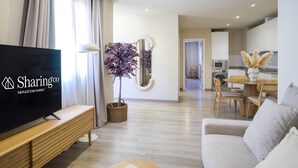 Un salon moderne avec un téléviseur à écran plat, un banc en bois, un canapé, un coin repas avec une table et des chaises, et une cuisine en arrière-plan.