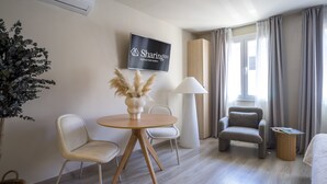 Una sala moderna con una mesa redonda de madera, dos sillas, un sofá, un televisor montado en la pared y una ventana con cortinas.