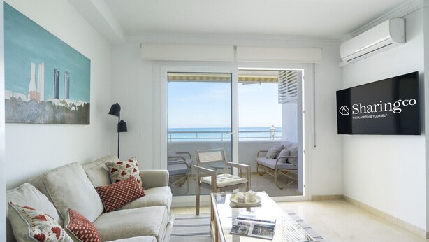 A modern living room with a sofa, coffee table, and a TV. There's a balcony with a view of the sea and buildings. - Amazing flat with views & pool by SharingCo  (Marbella)