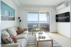 Ein modernes Wohnzimmer mit einer Couch, einem Couchtisch und einem Fernseher. Es gibt einen Balkon mit Blick auf das Meer und Gebäude.