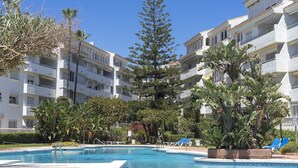 A pool area with a clear sky, surrounded by multi-story buildings with balconies, and various trees and plants. - Amazing flat with views & pool by SharingCo  (Marbella)