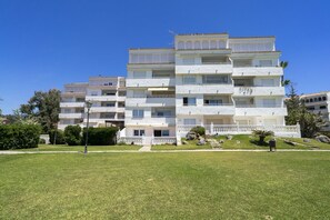 A multi-story residential building with balconies, a grassy front yard, and a clear blue sky.