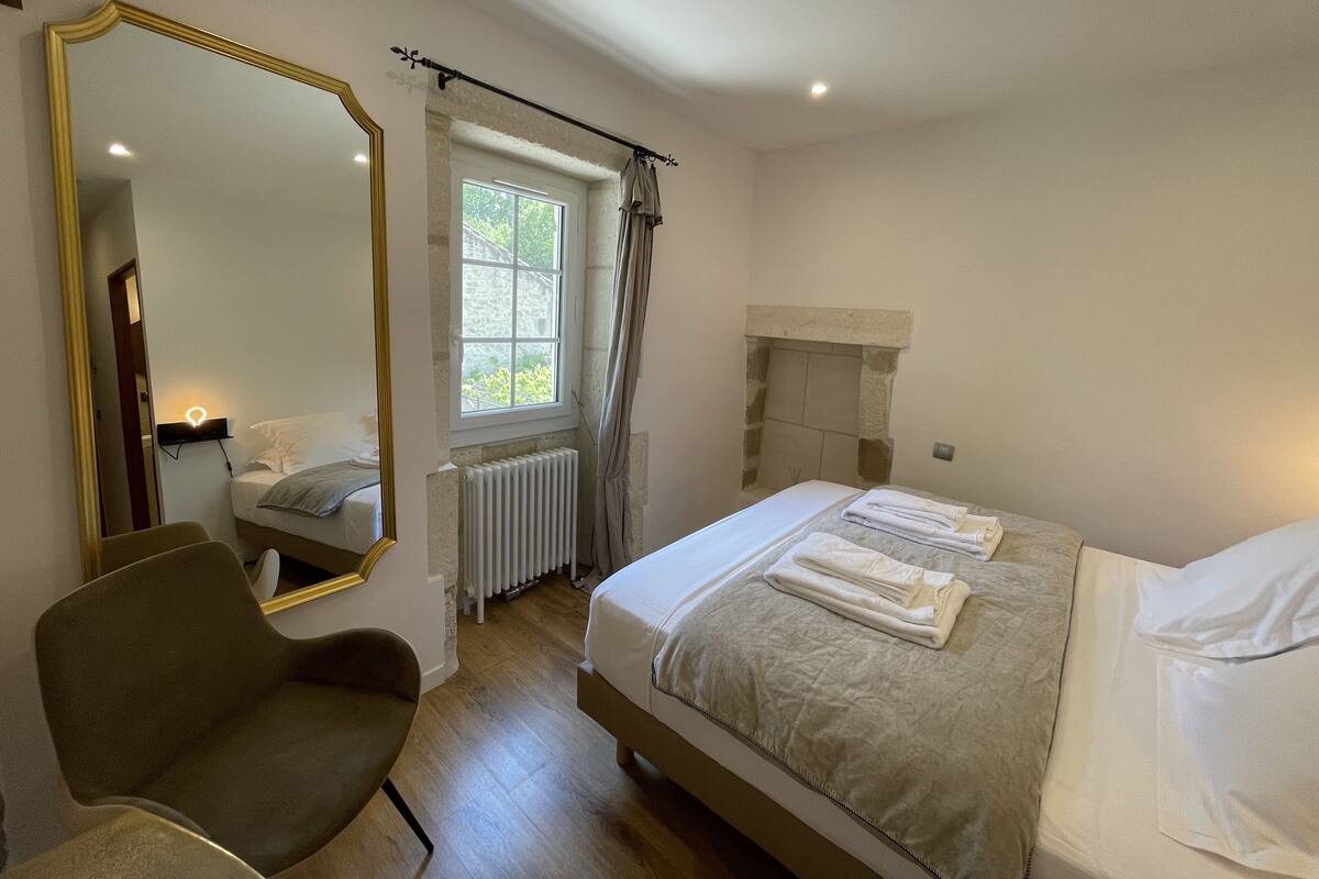 Un dormitorio con cama, sillón, espejo grande, ventana, radiador y chimenea.