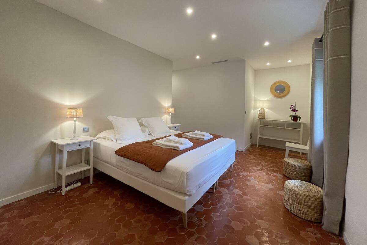 Habitación de hotel con una cama grande, dos mesitas de noche con lámparas, un pequeño rincón de descanso con un banco y un espejo en la pared.