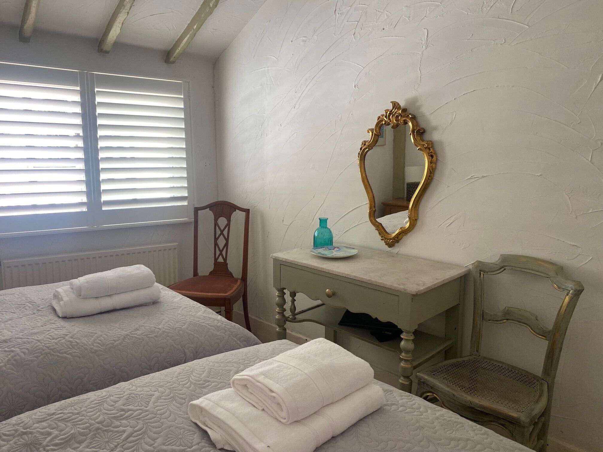 A room with two beds, a wooden chair, a small table with a mirror, and a window with blinds.