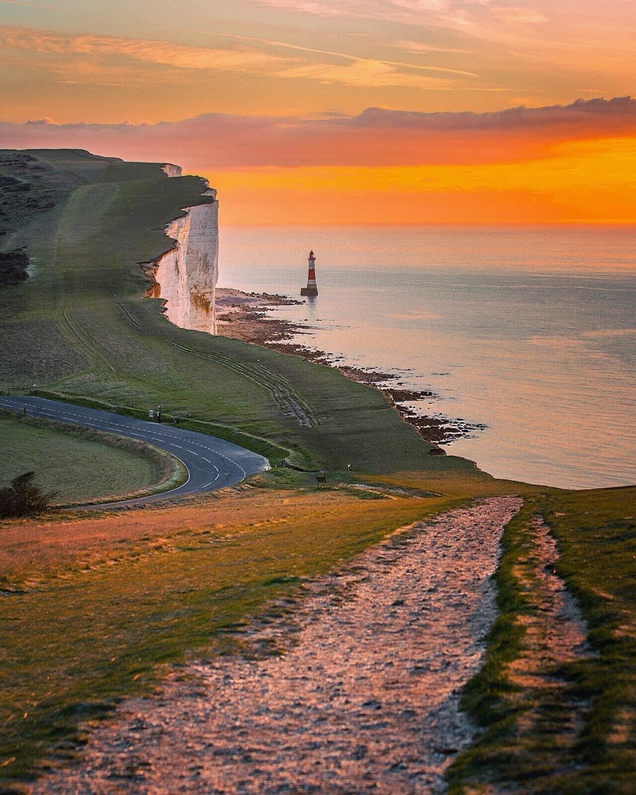 A coastal landscape with a road curving along the cliffs, a lighthouse, and a sunset over the sea.