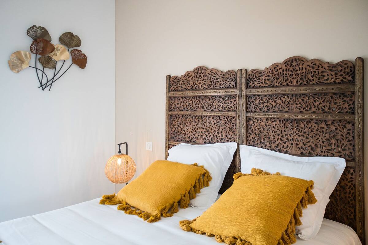 Una cama con sábanas blancas y dos almohadas color mostaza, un adorno en la pared y un panel de pared con textura.