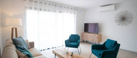 A modern living room with a sofa, two teal armchairs, a wooden coffee table, and a TV on a cabinet.