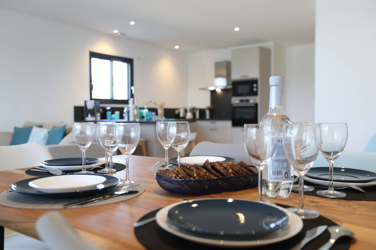Una mesa de comedor dispuesta con platos, cubiertos y copas de vino, con un bol de aperitivos en el centro.