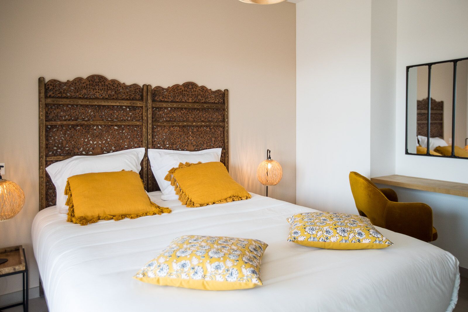 A neatly made bed with white linens and two mustard-colored pillows, a wooden headboard, and a bedside lamp.