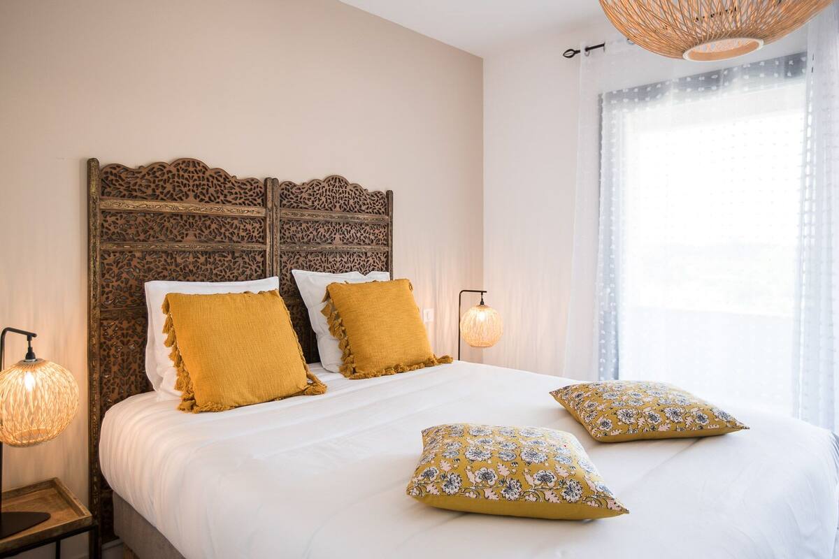 Un dormitorio con cama, dos almohadas, una mesita de noche, una lámpara y una ventana con cortinas.