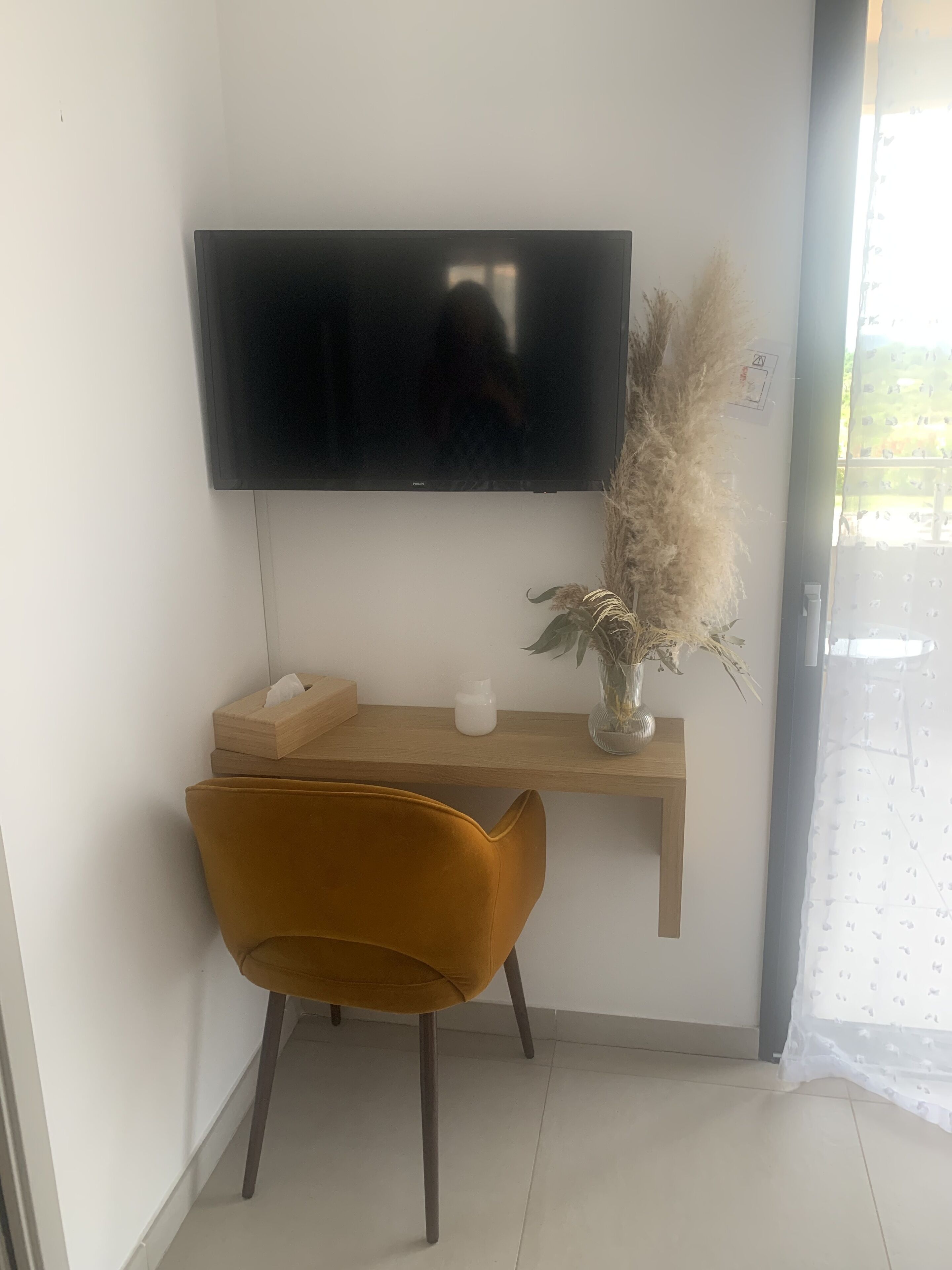 Una habitación moderna con un televisor montado, un estante de madera con objetos decorativos, una silla de color mostaza y un ventanal con vistas.