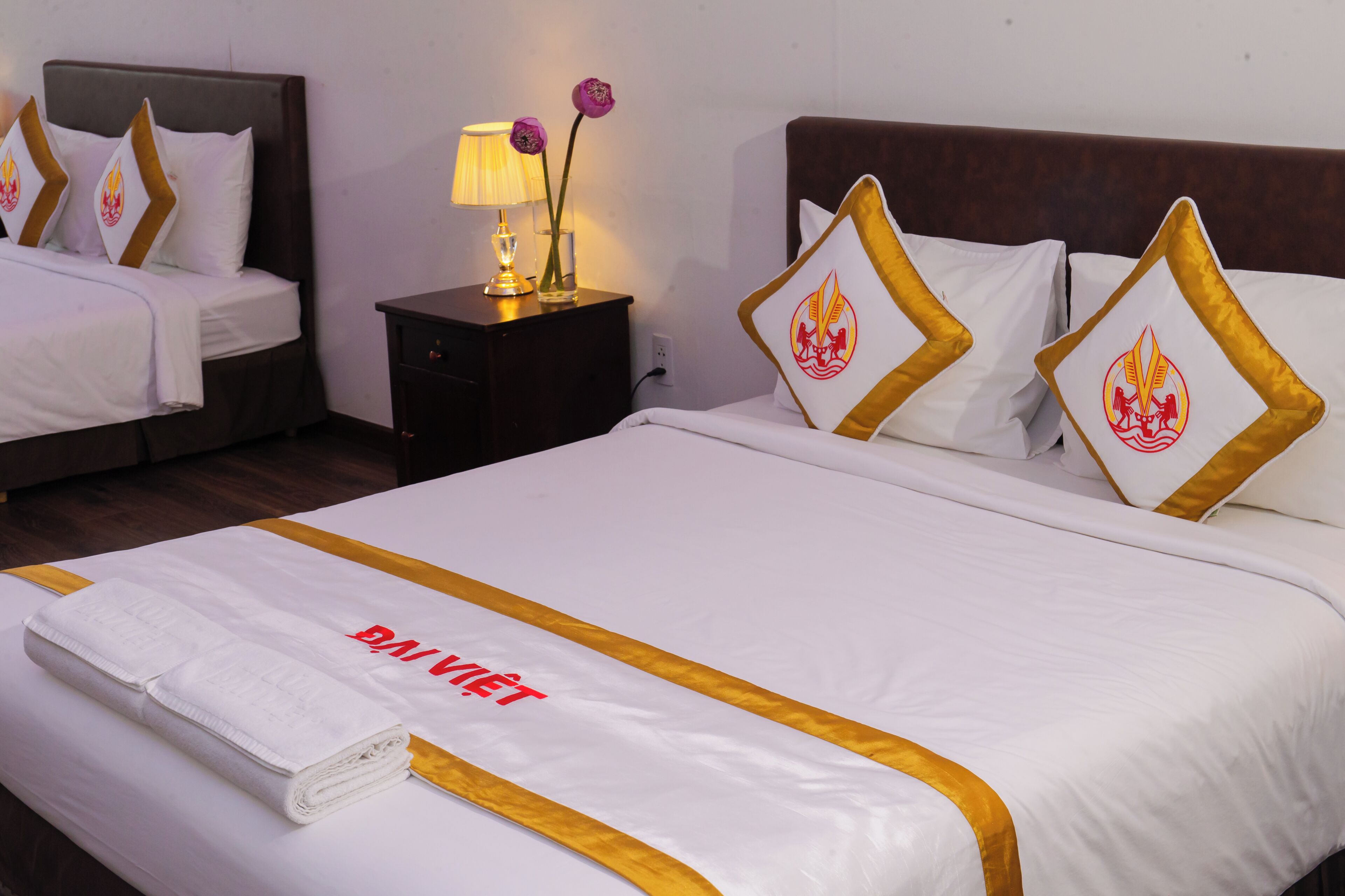Camera d'albergo con due letti, un comodino con lampada e fiori, e un letto rifatto con asciugamano e cartello.