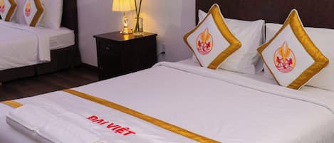 Camera d'albergo con due letti, un comodino con lampada e fiori, e un letto rifatto con asciugamano e cartello.