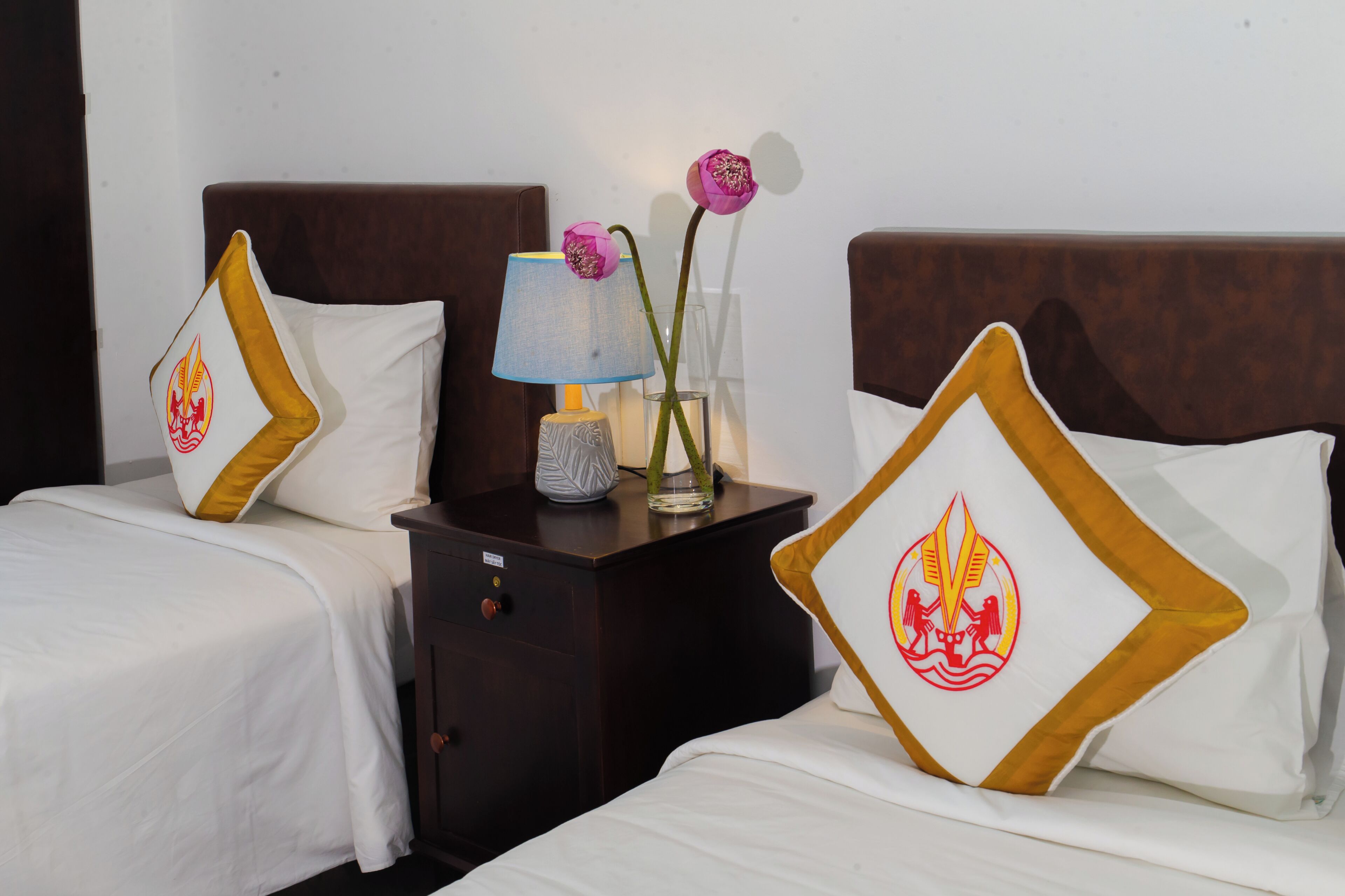 Une chambre d’hôtel avec deux lits, une table de chevet avec une lampe, un vase de fleurs et des coussins décoratifs.