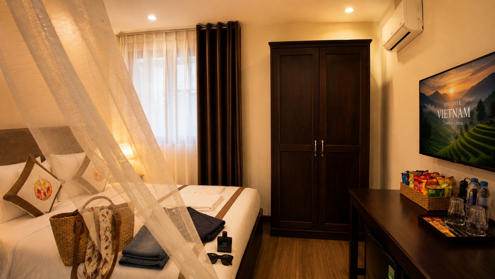 A hotel room with a bed, a wardrobe, a television, and a desk with a basket of snacks.