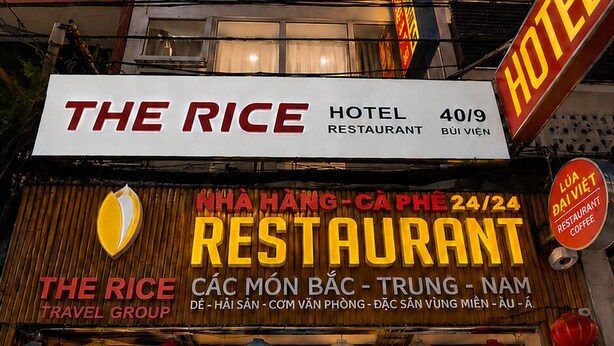 A hotel with a sign indicating it is 'The Rice Hotel Restaurant' and 'Nha Hương Café.' It has a reception area with a wooden counter and a large potted plant. - The Rice Hotel & Tour - 40 Bui Vien No 9 (Ho Chi Minh City)