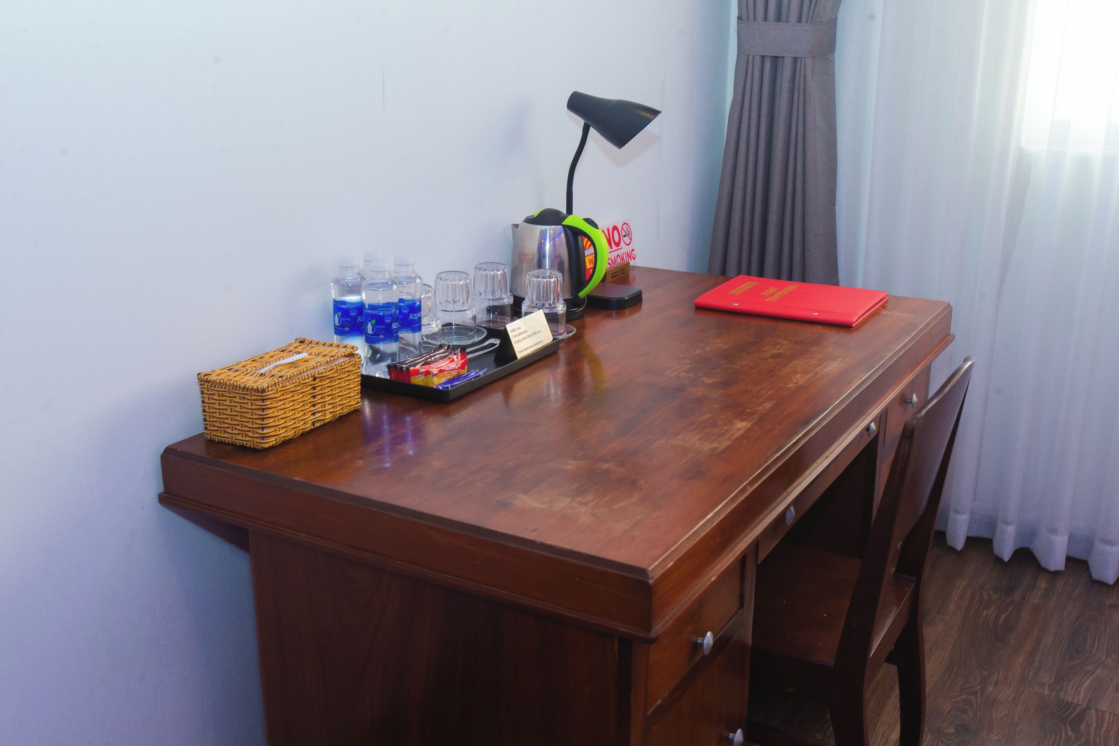A wooden desk with a lamp, a basket, a tray with glasses and bottles, and a red folder.