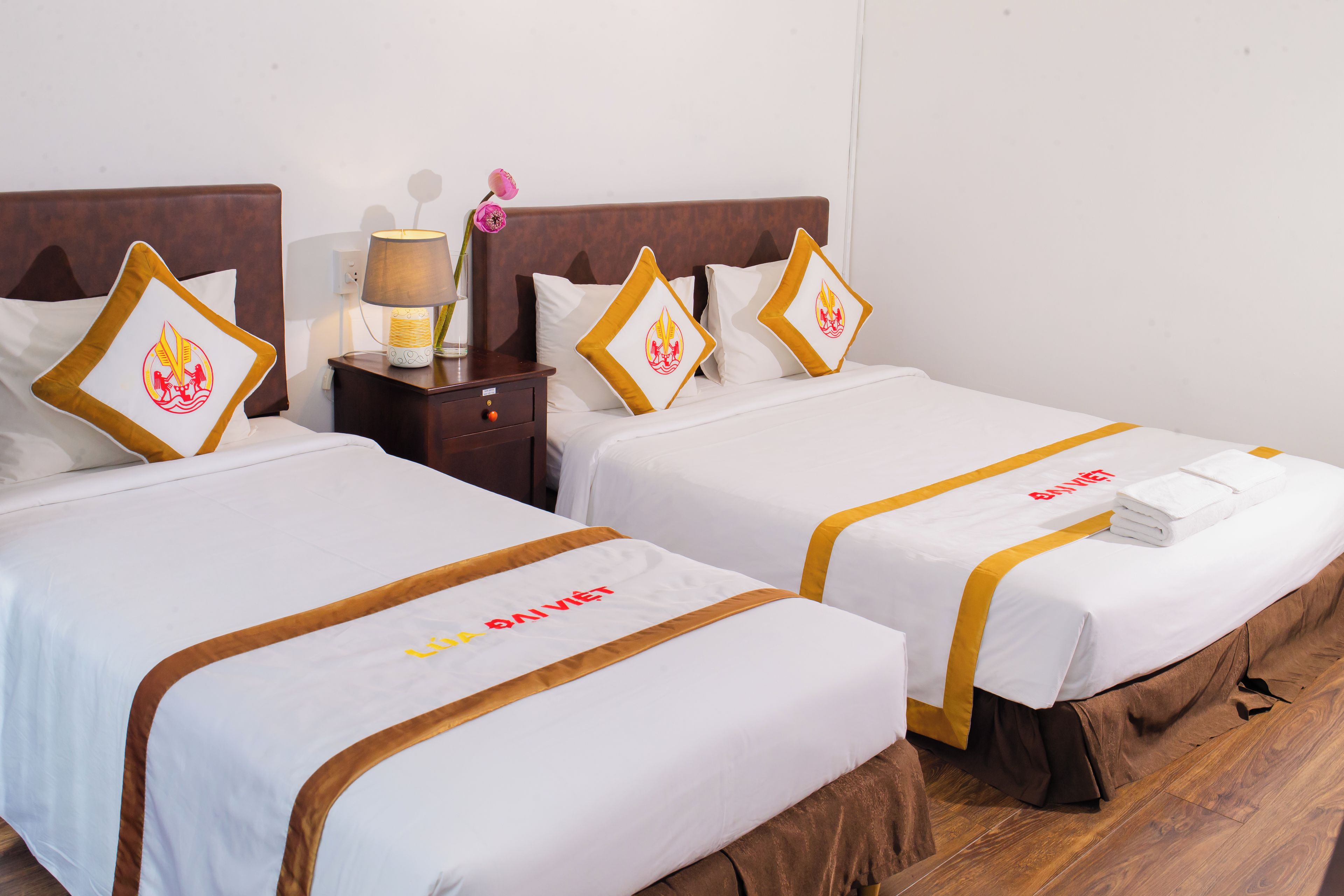 Habitación de hotel con dos camas, cada una con una colcha blanca y un borde dorado. Hay una mesita de noche con una lámpara y un florero con una flor entre las camas.