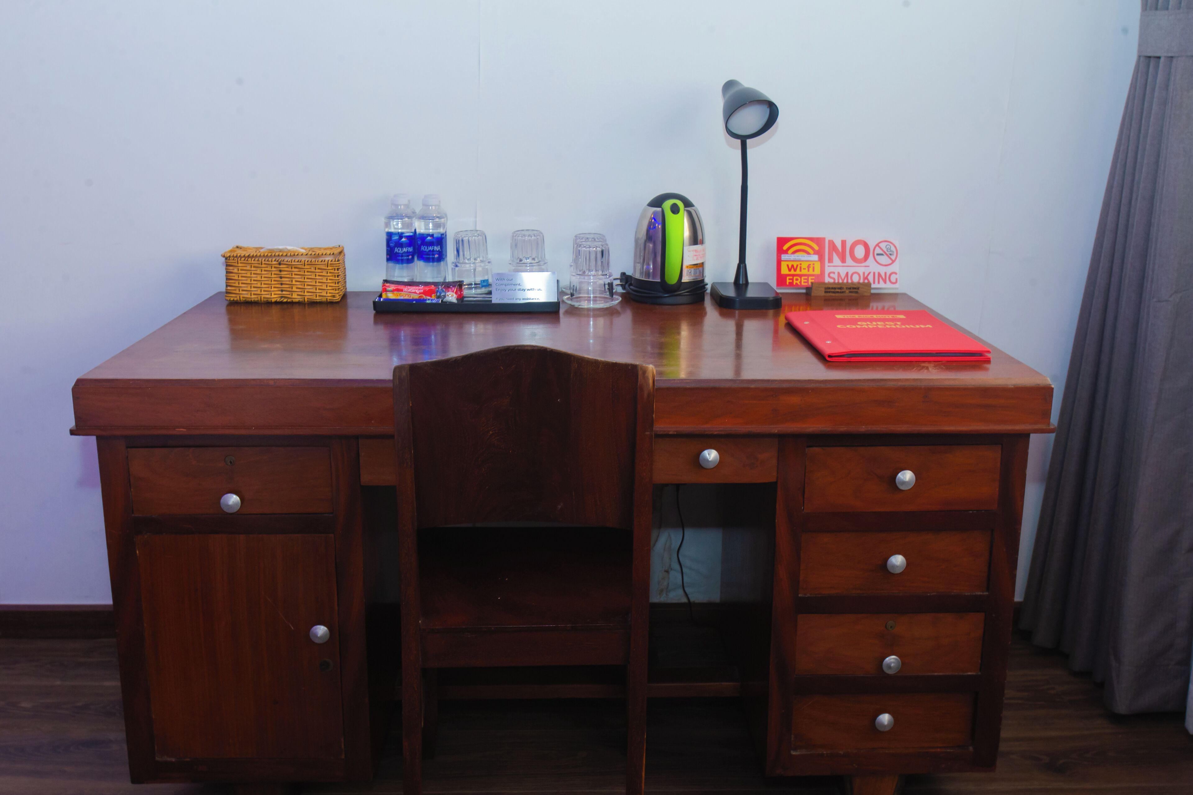 Un bureau en bois avec une chaise, un panier, une bouteille d’eau, une bouilloire, une lampe et un panneau indiquant « Il est interdit de fumer ».