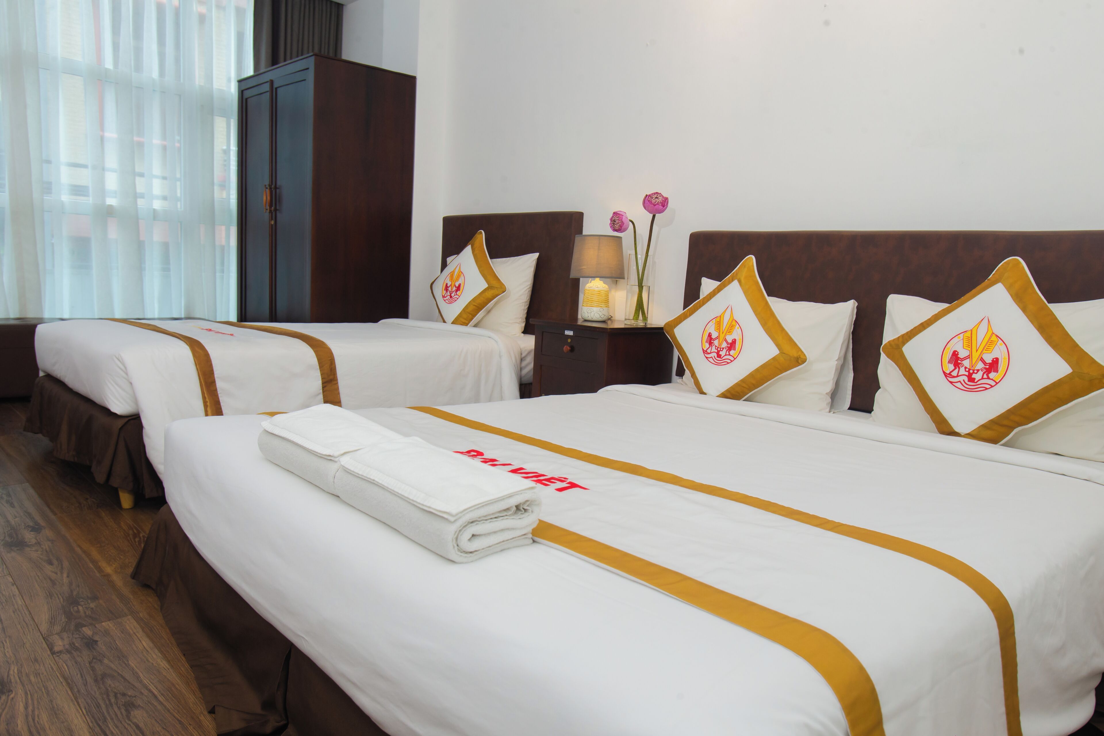 A hotel room with two beds, each with a white bedspread and gold trim, a nightstand with a lamp, and a wardrobe.